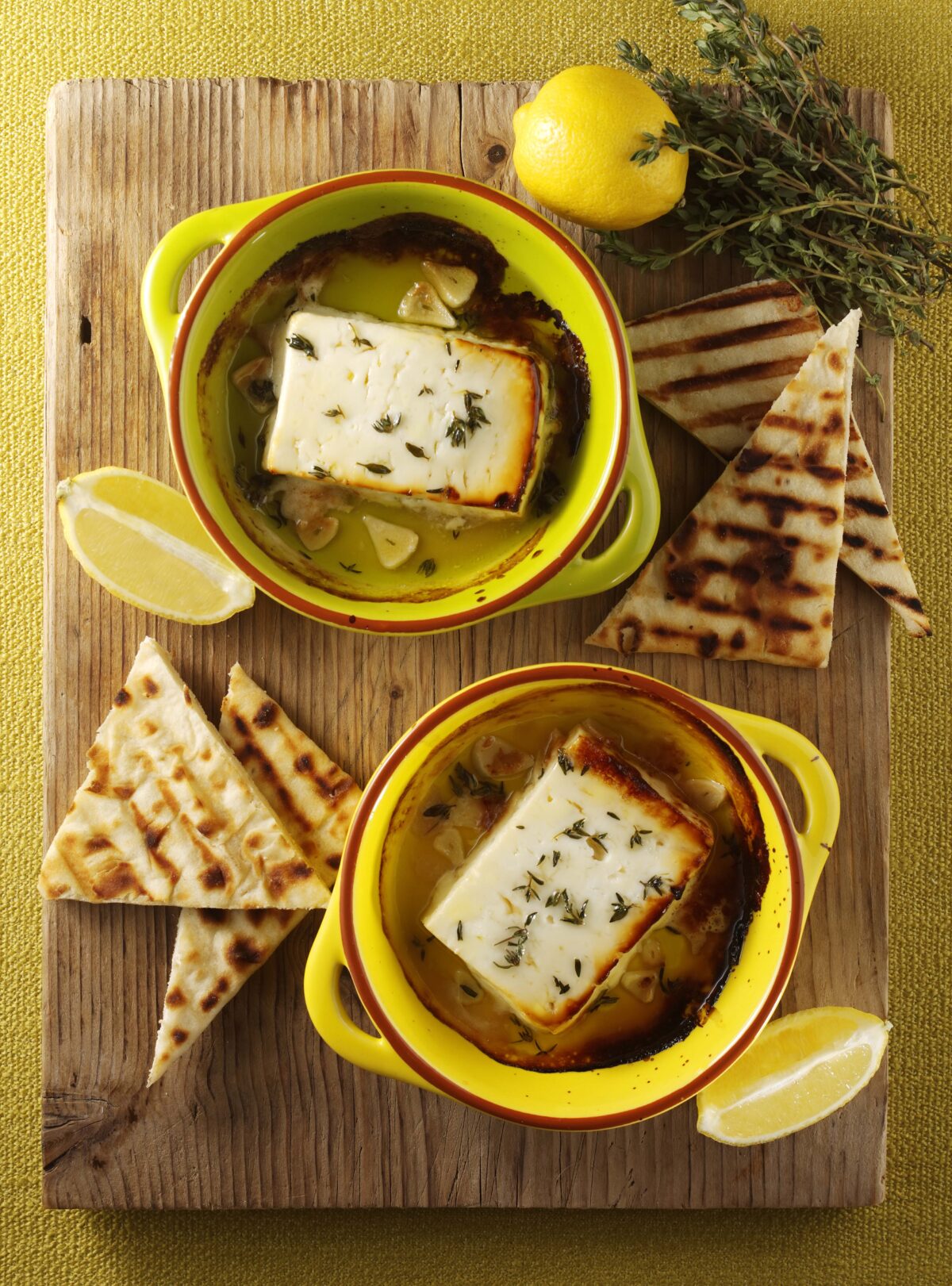 Two small, yellow dishes holding baked feta with lemon, garlic and herbs on a chopping board next to toasted bread and lemons