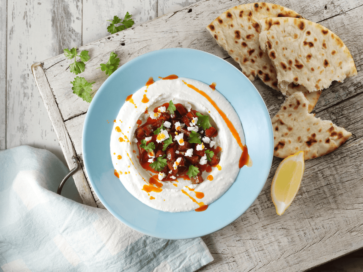 Whipped feta and chorizo in a blue dish on a wooden board next to lemon wedge, bread, herbs and a napkin