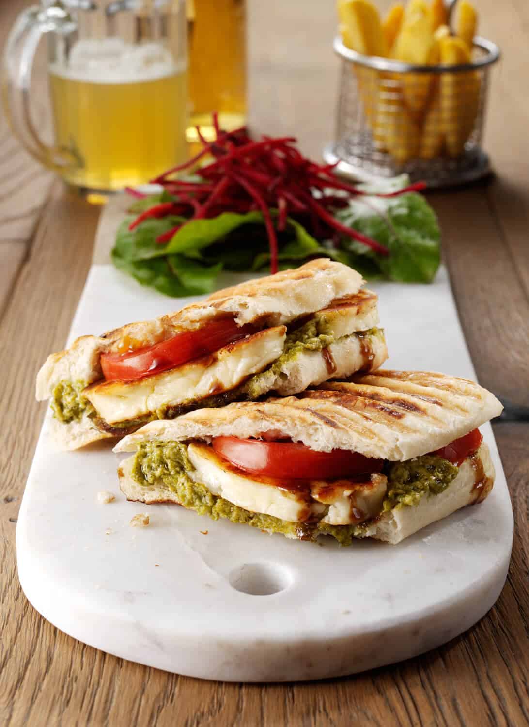 A toasted panini filled with a layer of pesto adn slices of halloumi and tomatoes. On a white marble serving board with a side of salad.