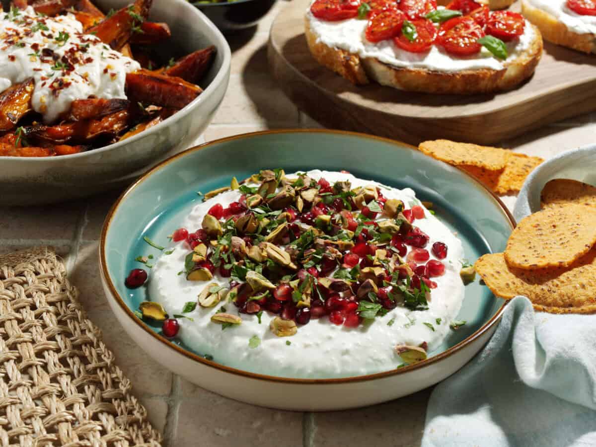 A shallow bowl filled with creamy YAMAS! Whipped Feta, topped with vibrant pomegranate seeds, chopped pistachios, and fresh herbs, served with golden pita chips on a wooden surface.