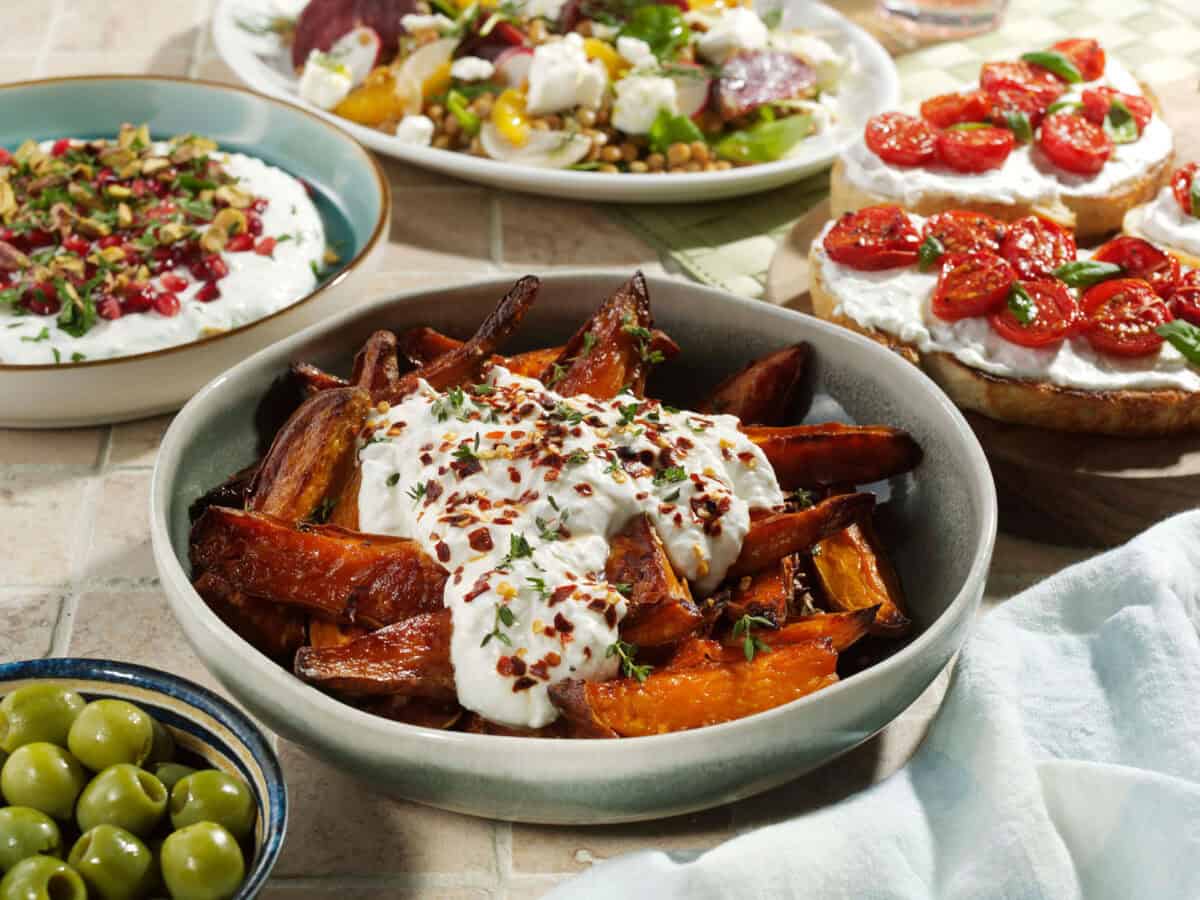 A ceramic bowl filled with golden-brown roasted sweet potato wedges topped with a generous layer of YAMAS Whipped Feta, sprinkled with fresh thyme and chilli flakes, presented on a rustic surface.