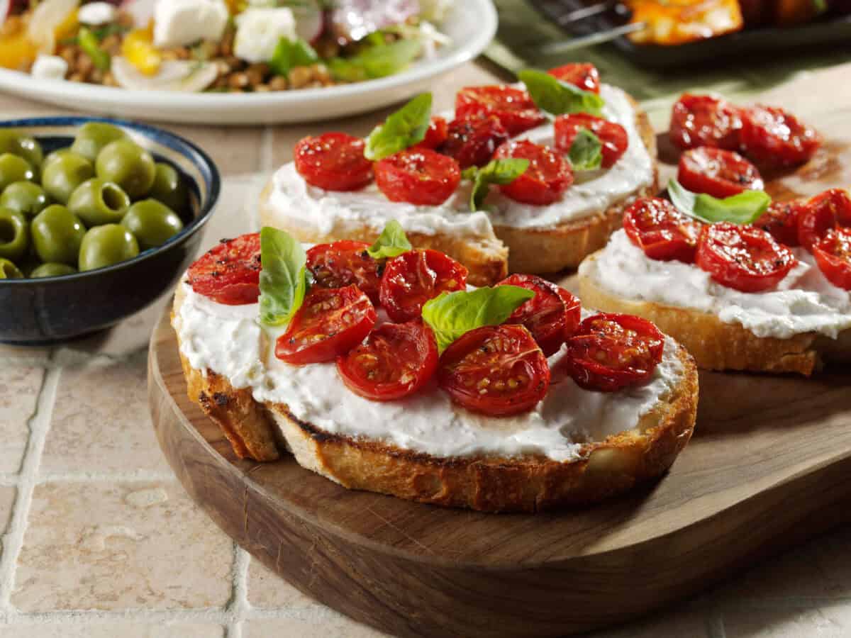 Two slices of crusty sourdough bread topped with creamy YAMAS! Whipped Feta, roasted blistered cherry tomatoes, and fresh basil, served on a wooden board next to a bowl of green olives.